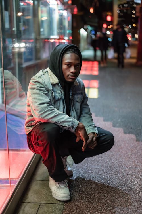 Free Full body of serious African American male in hood looking at camera while sitting on haunches near glass wall in evening time Stock Photo