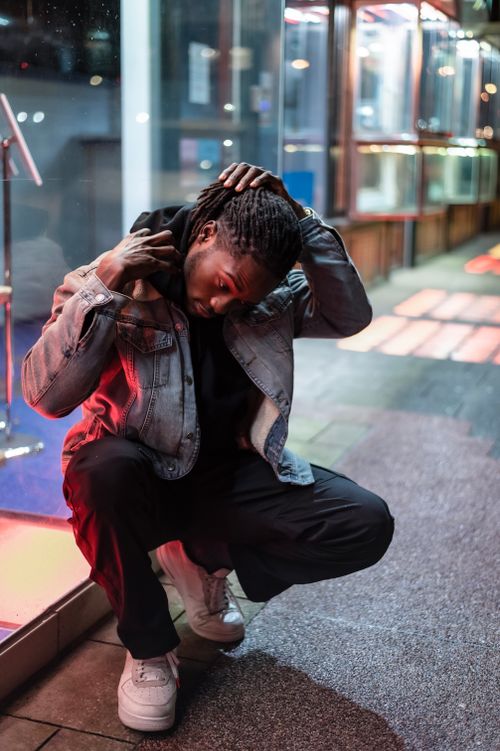 Free Full body of confident African American male with dreadlocks wearing denim jacket hunkering down on empty walkway near building on evening time Stock Photo