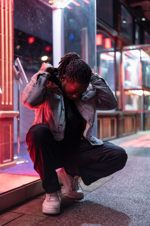 Free Full body of young African American male with dreadlocks sitting on haunches on street and putting on hood against luminous lights at night Stock Photo