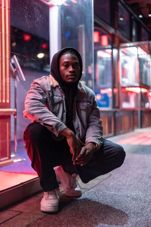 Free Full body of serious African American male in hood sitting on sidewalk against bright lights at night Stock Photo