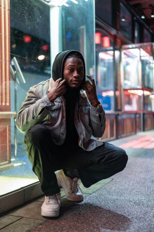 Free Full body of self assured young African American male in hood of sweatshirt sitting on sidewalk near glass wall and looking at camera Stock Photo