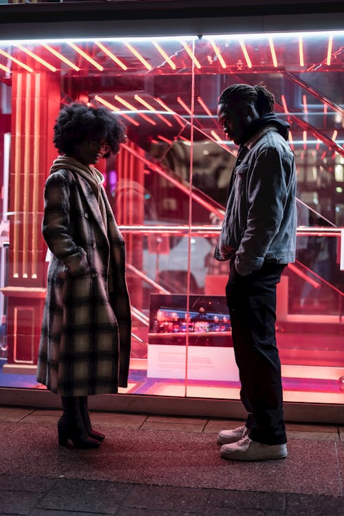 Free Side view full body of offended African American couple standing with hands in pockets near glowing lights behind glass wall Stock Photo