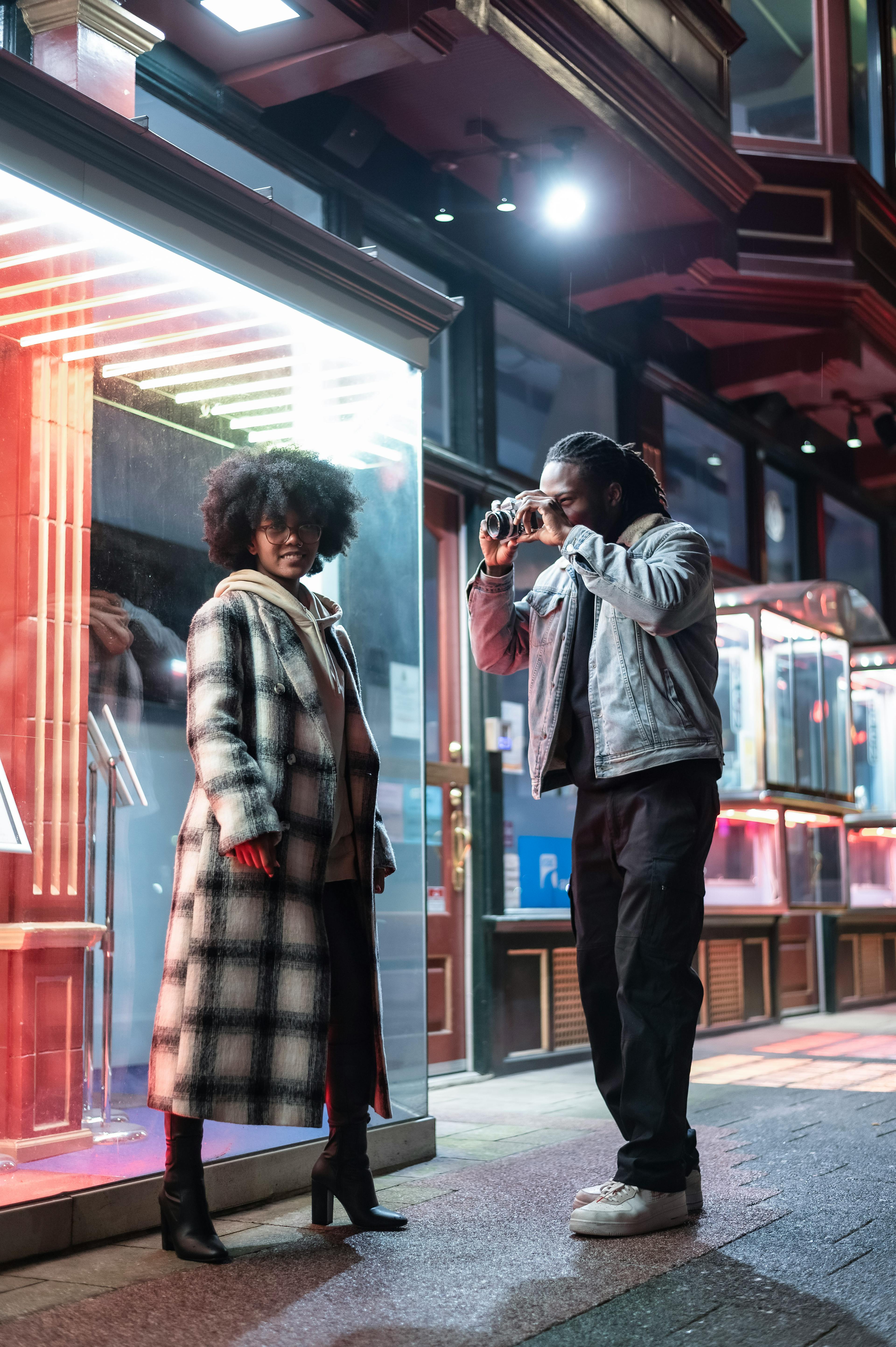 Full body of focused African American male photographer taking picture of positive black female looking at camera while standing on paved sidewalk in evening time