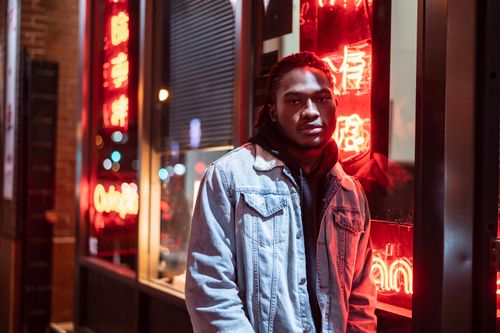 Free African American male wearing casual clothes leaning on glass wall with glowing red neon illumination and looking at camera Stock Photo