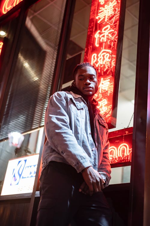 Low angle of thoughtful African American male wearing casual outfit leaning on wall with glowing red neon illumination