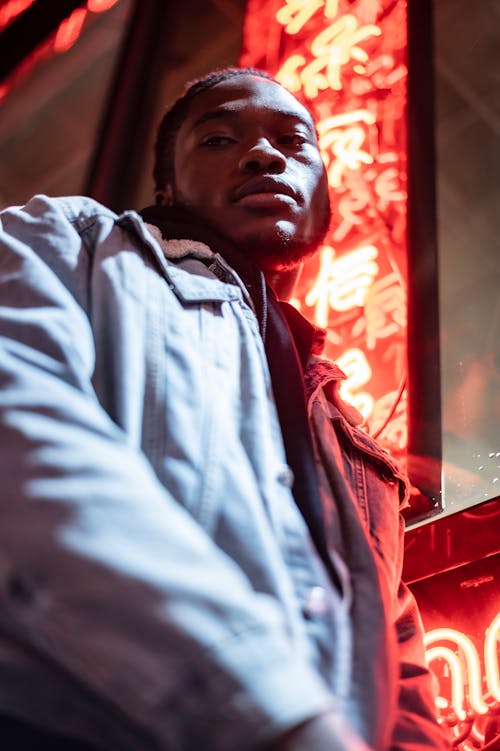 Low angle of African American male in denim jacket standing near building with neon hieroglyphs