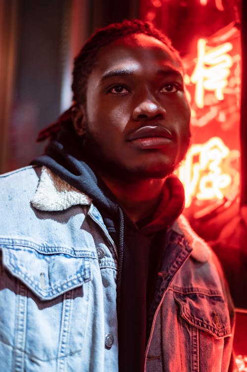 Dreamy African American male in denim jacket and hoodie against luminous red neon lights at night