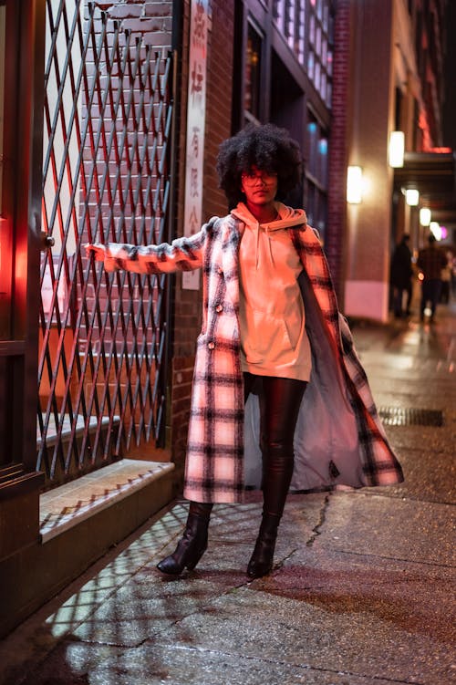 Full body of African American female in eyeglasses and coat on hoodie standing on wet sidewalk at night