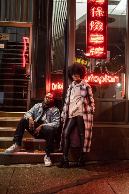 Stylish ethnic woman in checkered coat against man on staircase looking at camera in city at dusk