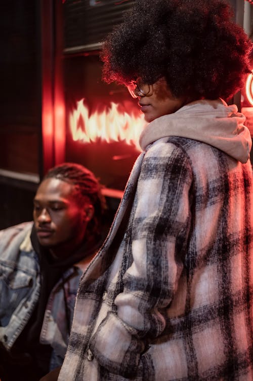 Stylish ethnic woman with hand in checkered coat pocket and Afro hairstyle against man in evening city