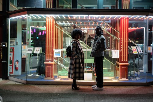 Side view of trendy black woman in checkered coat against man with hands in pockets on pavement at dusk