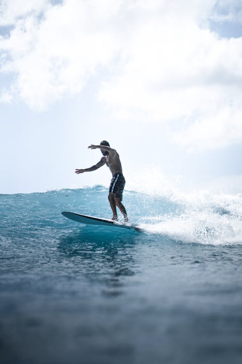 Full body of shirtless anonymous male surfer standing on surfboard in wavy sea under blue cloudy sky in daytime