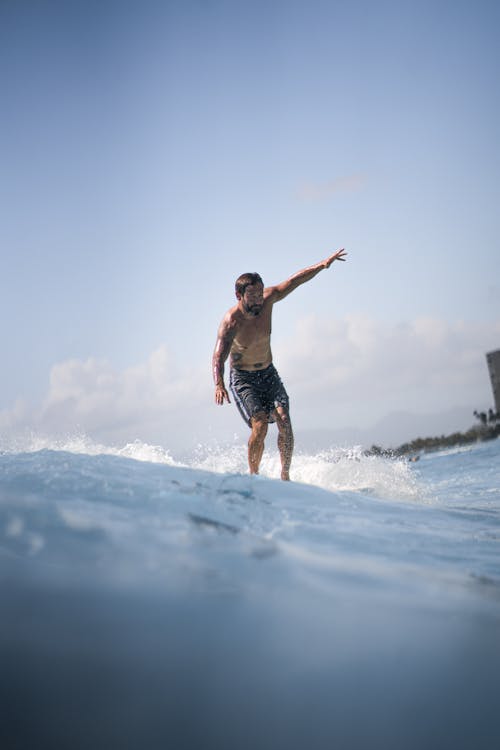 Full body of shirtless male surfer in shorts standing on surfboard in wavy sea against cloudy sky