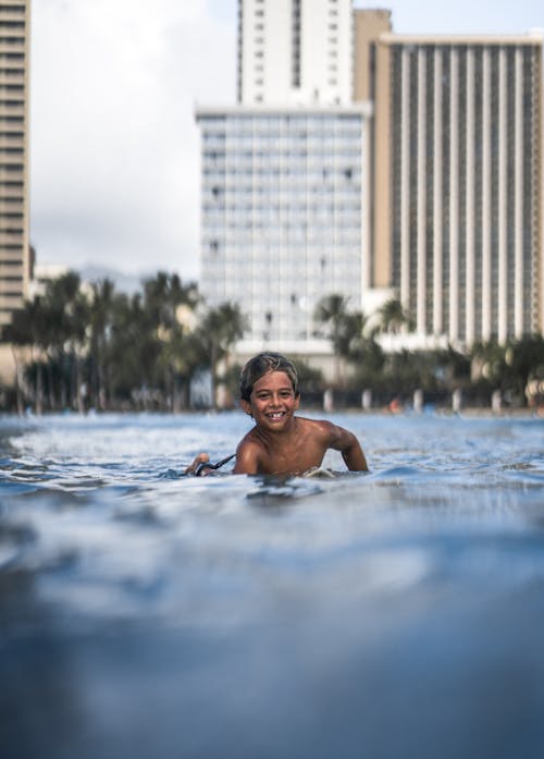 Free Smiling ethnic child swimming in open pool with rippling water at resort in summer on blurred background of modern city with multistory buildings Stock Photo