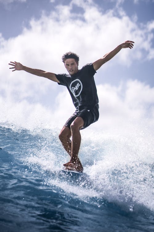 Low angle of young man in t shit and shorts spreading arms apart while surfing splashing wave in sea