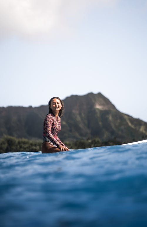 Free Happy female in swimsuit sitting on surfing board in clear ocean water and looking at camera against mountains Stock Photo