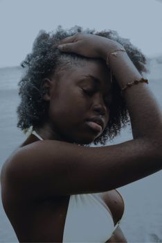 A serene Afro-American woman with curly hair poses by the sea wearing a white bikini top.
