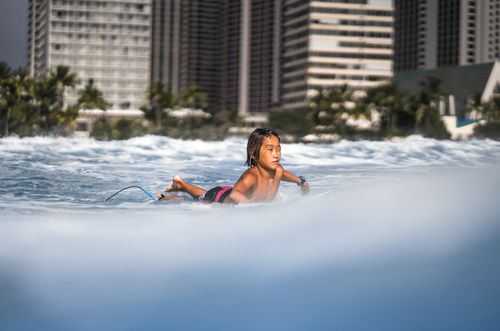 Free Shirtless Asian boy lying on surfboard while swimming in sea on sunny day against blurred background of city Stock Photo