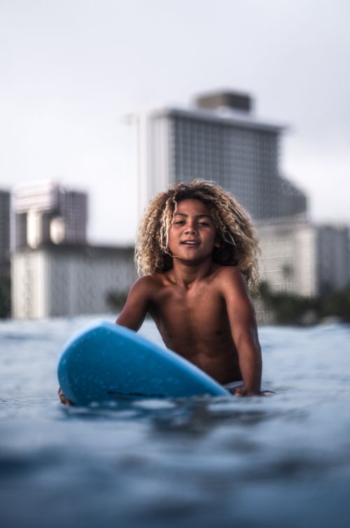 Free Fair haired boy sitting on surfboard in sea water and looking at camera on blurred background of cityscape Stock Photo