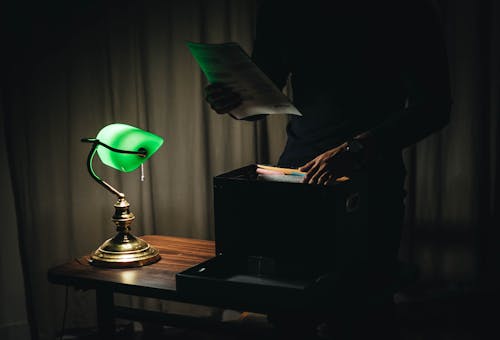 Unrecognizable male with sheets of paper standing near wooden table with glowing table lamp and box of documents in dark room while working late at home