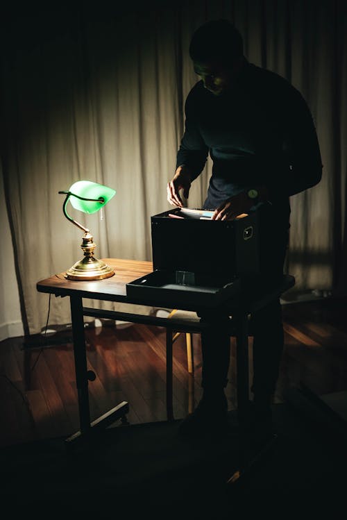 Unrecognizable male standing at table and organizing papers in black box while working in room with glowing vintage table lamp at home