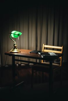 Chair at wooden desktop with glowing old fashioned lamp placed near black notepad and various documents in dark room with curtains