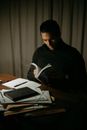 Concentrated male turning pages of book while sitting at wooden table with document during work at home in obscure room