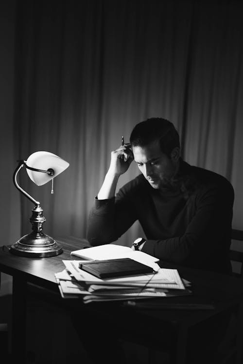 Free Black and white of concentrated male with pen doing paperwork while sitting at desktop with papers in room during work at home Stock Photo