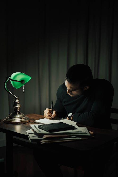 Concentrated male taking notes in notepad while sitting at desktop with documents in dark room with table lamp during paperwork at home