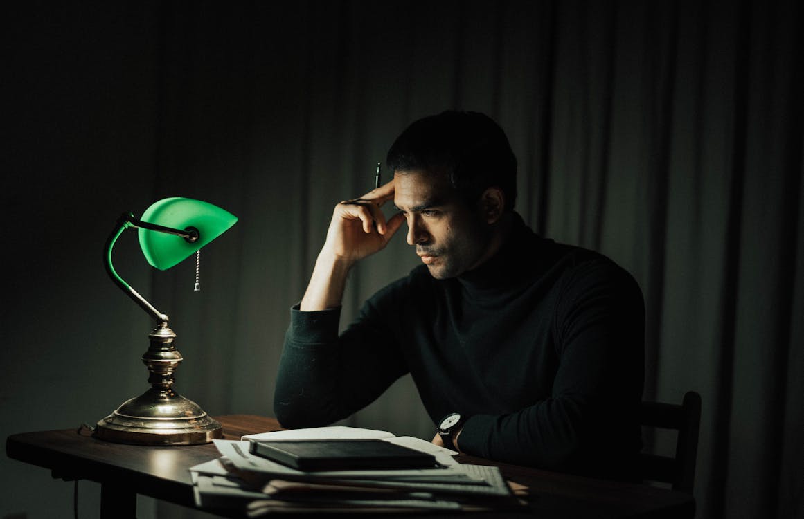 Side view of pensive male with pen doing paperwork while sitting at wooden desktop with vintage table lamp during work in obscure room