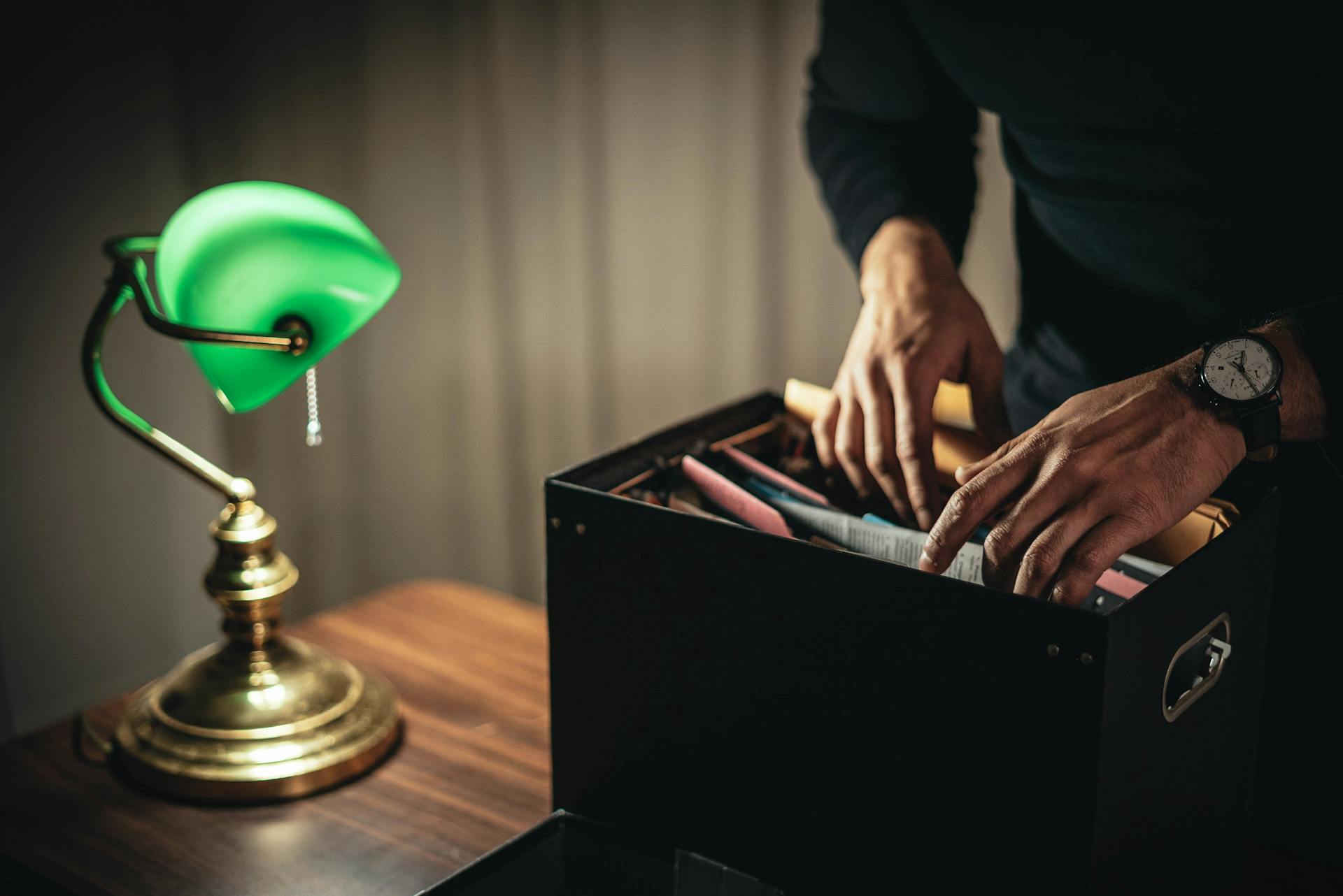 Crop anonymous male looking through files and folder in box placed on desk with green bankers lamp in home office