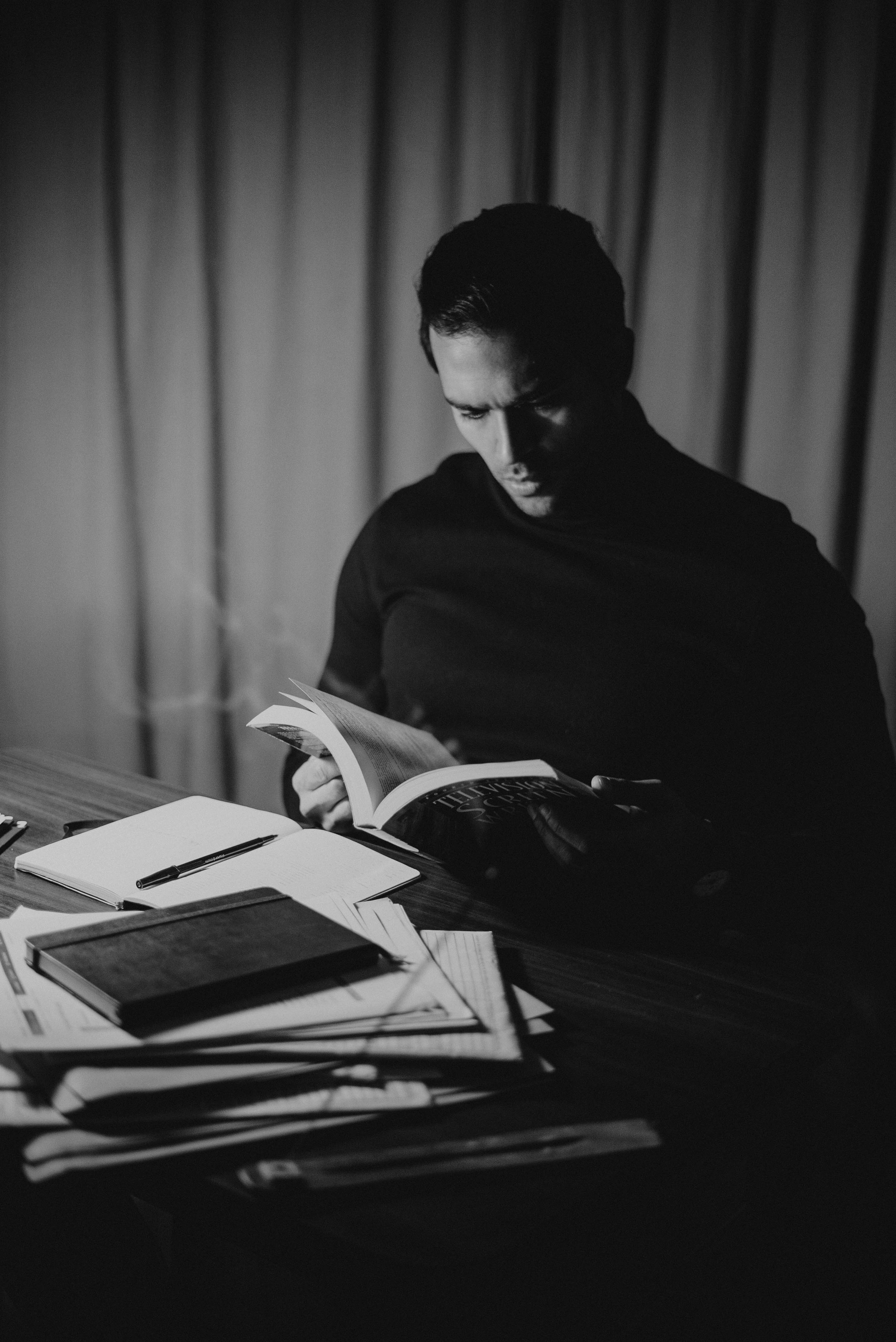 Black and white concentrated handsome male in dark turtleneck reading diary attentively while sitting at desk with papers in home office