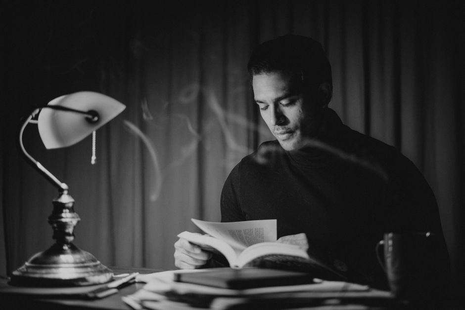 Black and white focused good looking male in casual wear reading book while sitting at desk with papers and bankers lamp in home office Black and white focused good looking male in casual wear reading book while sitting at desk with papers and bankers lamp in home office