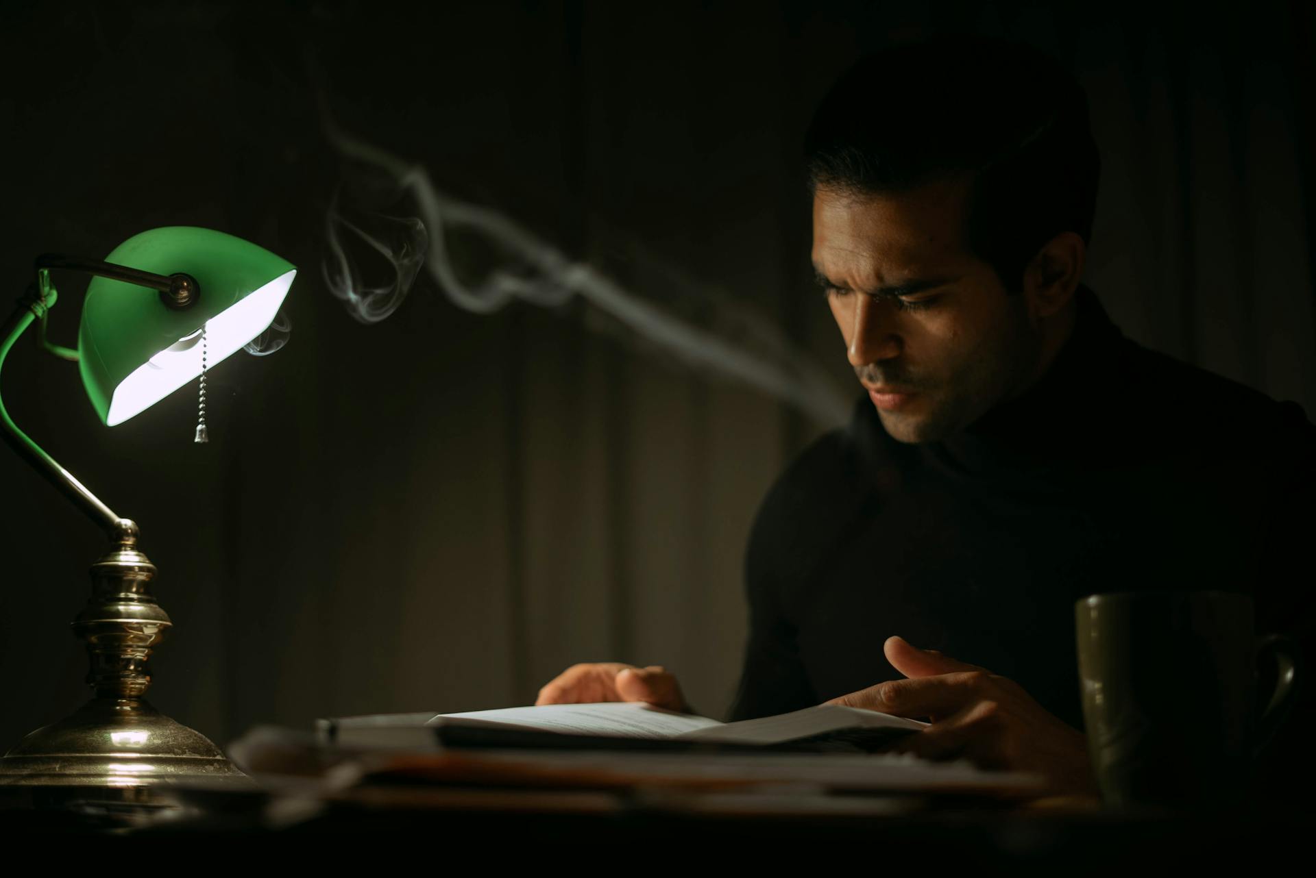 Pondering young male in black sweater reading book while sitting at desk with papers and green bankers lamp in dark home office