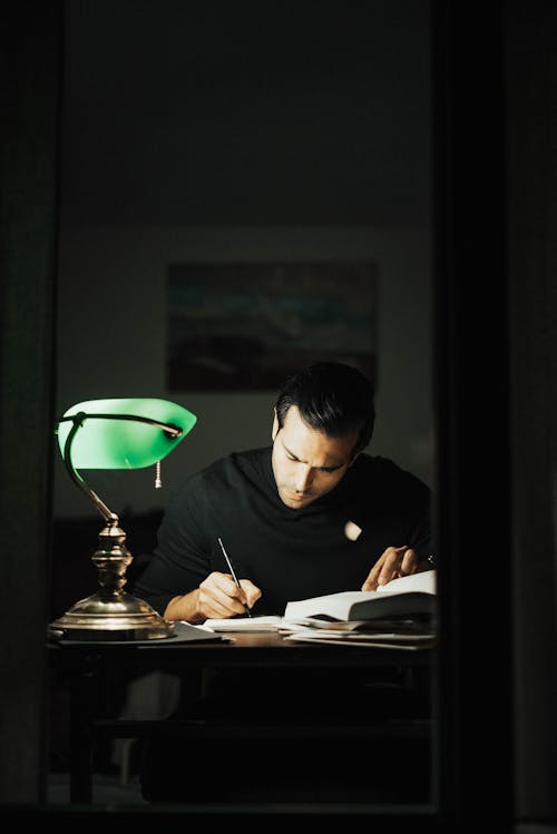 Concentrated young male wearing black turtleneck taking notes and reading books while sitting at desk with papers and bankers lamp in dark home office