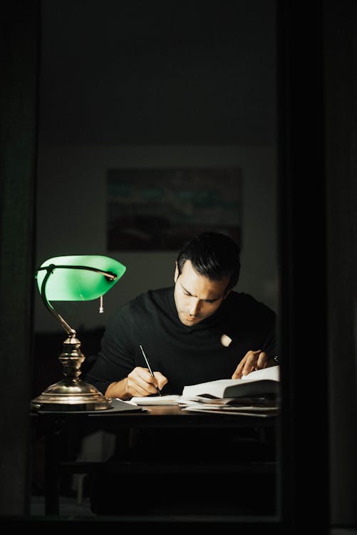 Free Concentrated young male wearing black turtleneck taking notes and reading books while sitting at desk with papers and bankers lamp in dark home office Stock Photo