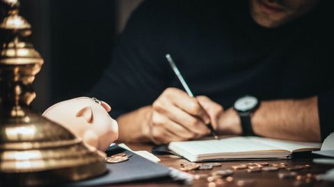 Crop man sitting at desk with vintage lamp and scattered coins and piggybank while writing in notepad
