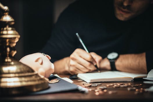A man writing in a notebook at a desk with a piggy bank and scattered coins, suggesting financial planning.