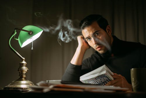 Handsome mature man in black shirt sitting at table with green lamp holding book in smoke and looking at camera