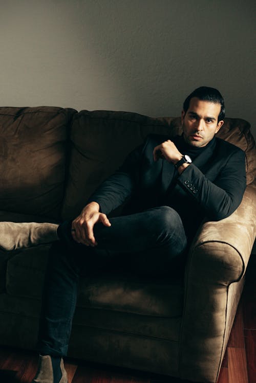 Full body adult serious man in posh suit sitting confidently on sofa and looking at camera