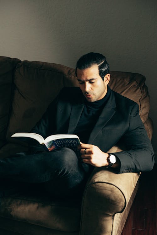 High angle of classy man wearing elegant black suit while sitting on sofa in light and reading book