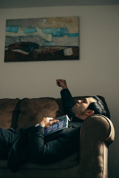 Side view of masculine handsome male in black suit lying on sofa with book and looking up