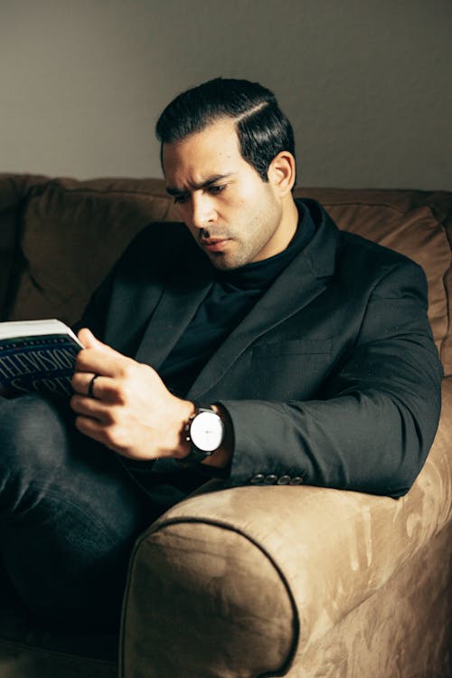 Confident classy guy in black suit sitting on couch and reading book in dim light