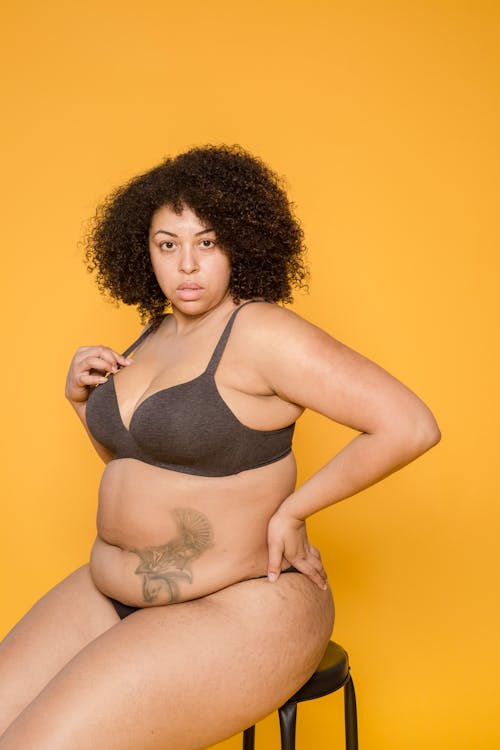 Obese African American female in bra and bikini sitting with hand on hip while looking at camera on yellow background