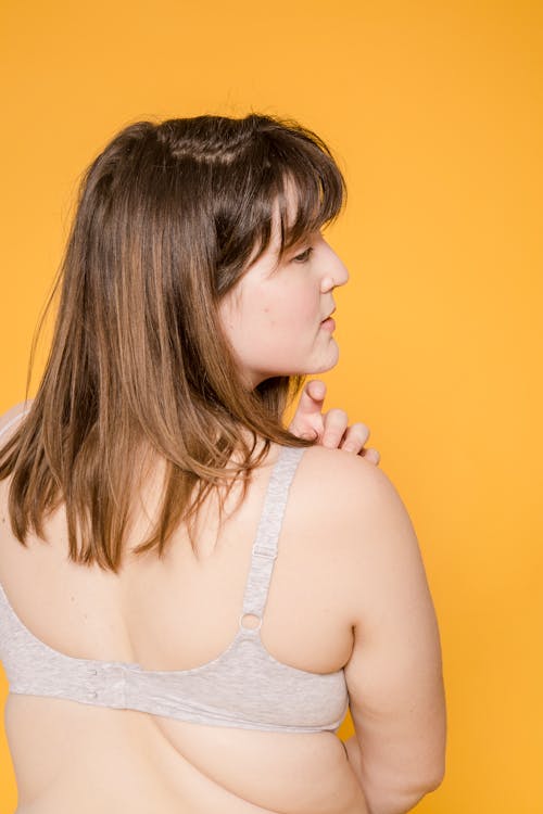 Back view of young plus size Asian woman with brown hair in bra looking away on yellow background