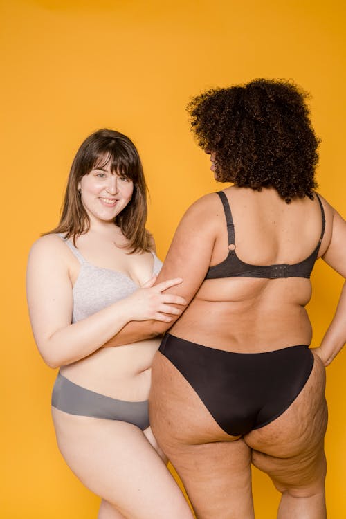 Smiling plus size Asian woman looking at camera near unrecognizable ethnic girlfriend with hand on hip and Afro hairstyle