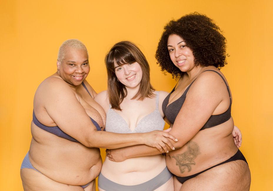 Happy overweight multiethnic women with tattoo and skin folds in underwear embracing while looking at camera