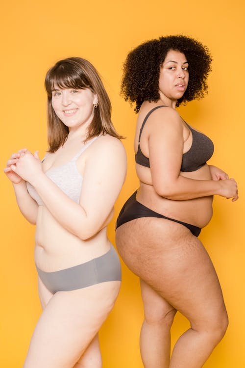 Cheerful plump Asian woman and plus size African American female in lingerie standing together on yellow background in light studio