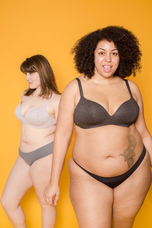 Free Positive overweight African American female in underwear looking at camera near plum Asian woman in lingerie on yellow background in studio Stock Photo