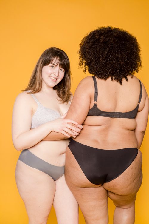 Free Cheerful overweight Asian woman in underwear looking at camera while standing with faceless plus size African American female on yellow background Stock Photo